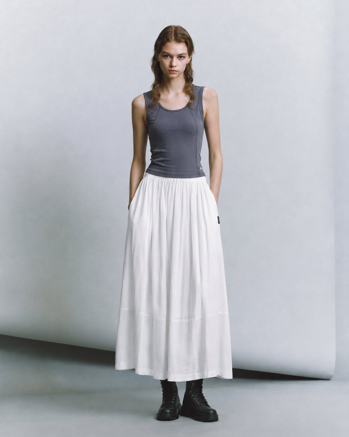A model stands in a studio wearing a dark gray sleeveless top, a long white skirt, and black boots against a light gray backdrop.